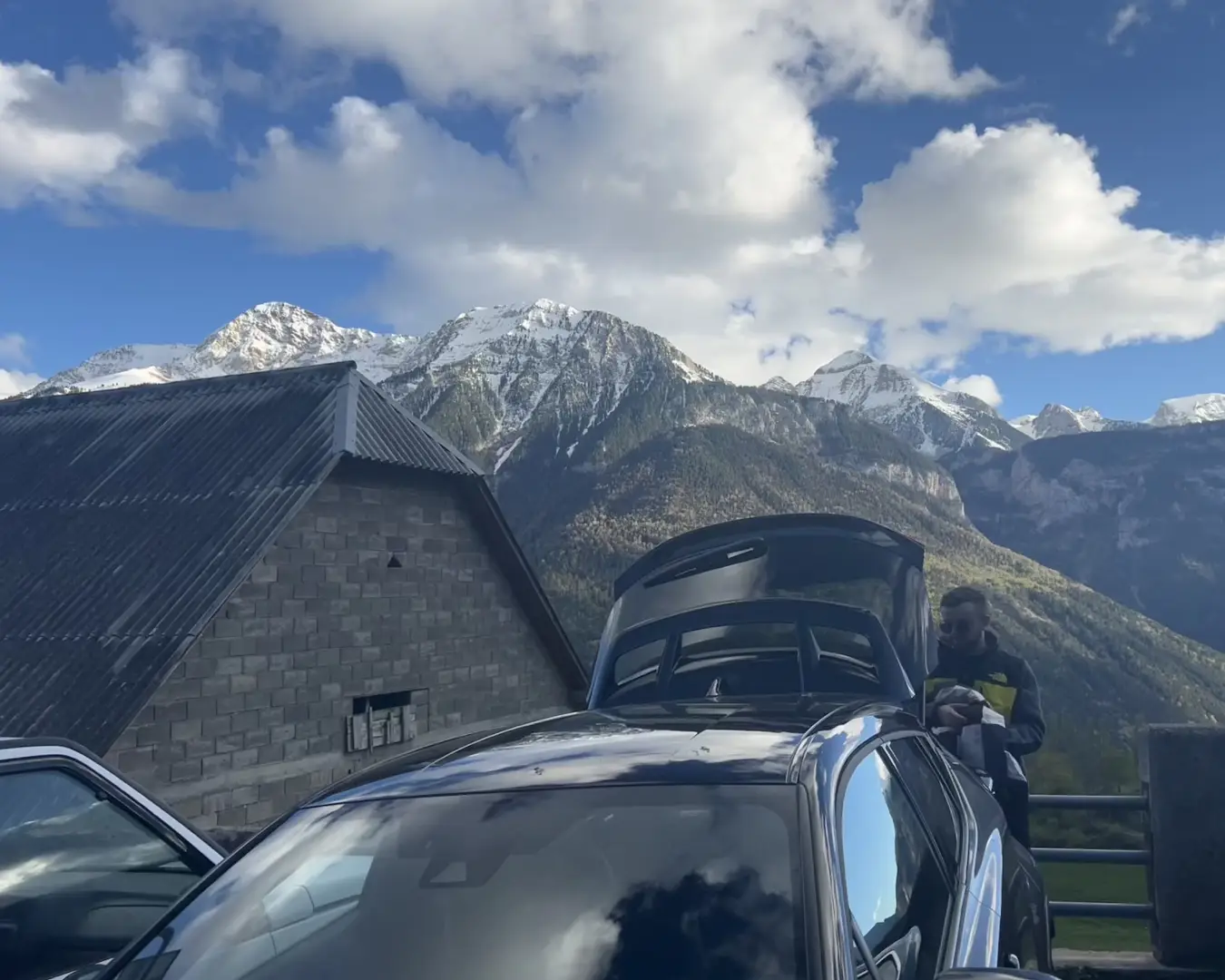Vida Cotidiana en los Pirineos: Montañas y Tradiciones Escena cultural en los Pirineos con montañas, una persona descargando un coche y una casa típica.